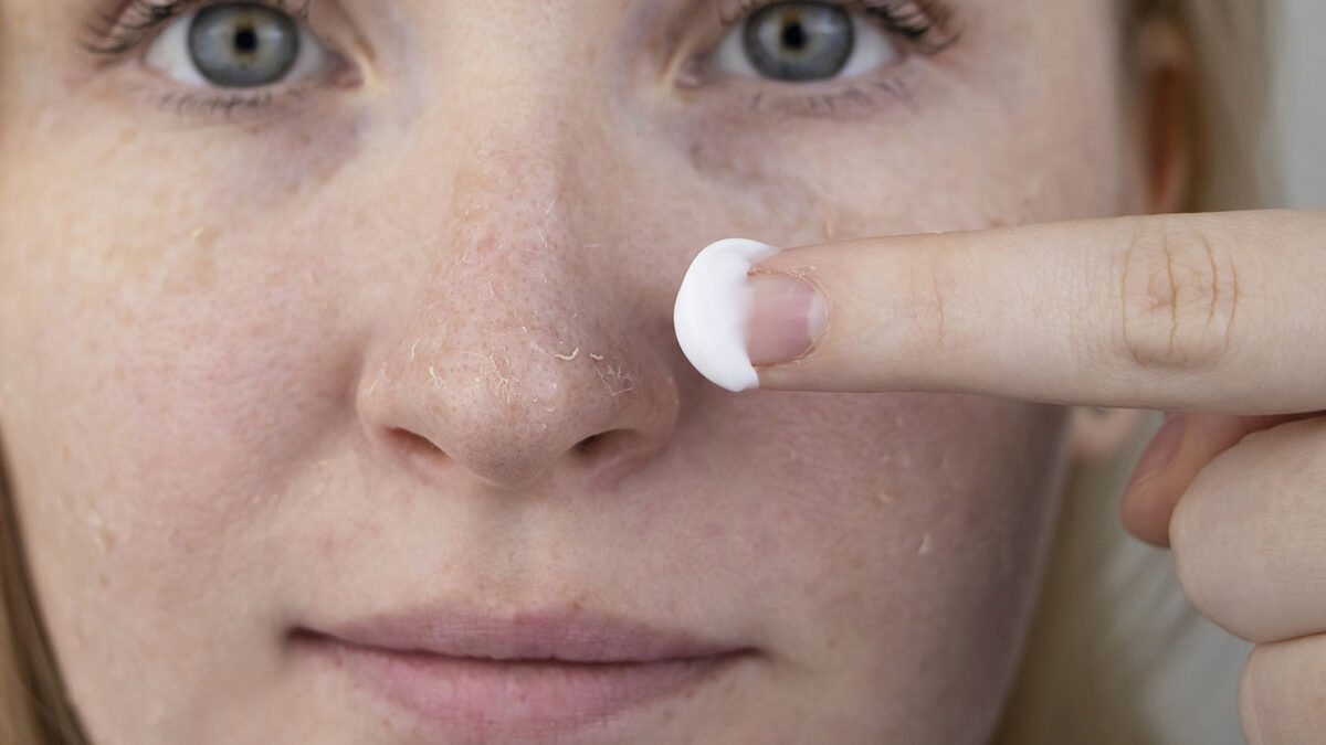 Uma mulher examina a pele seca em seu rosto e passa hidratante no nariz