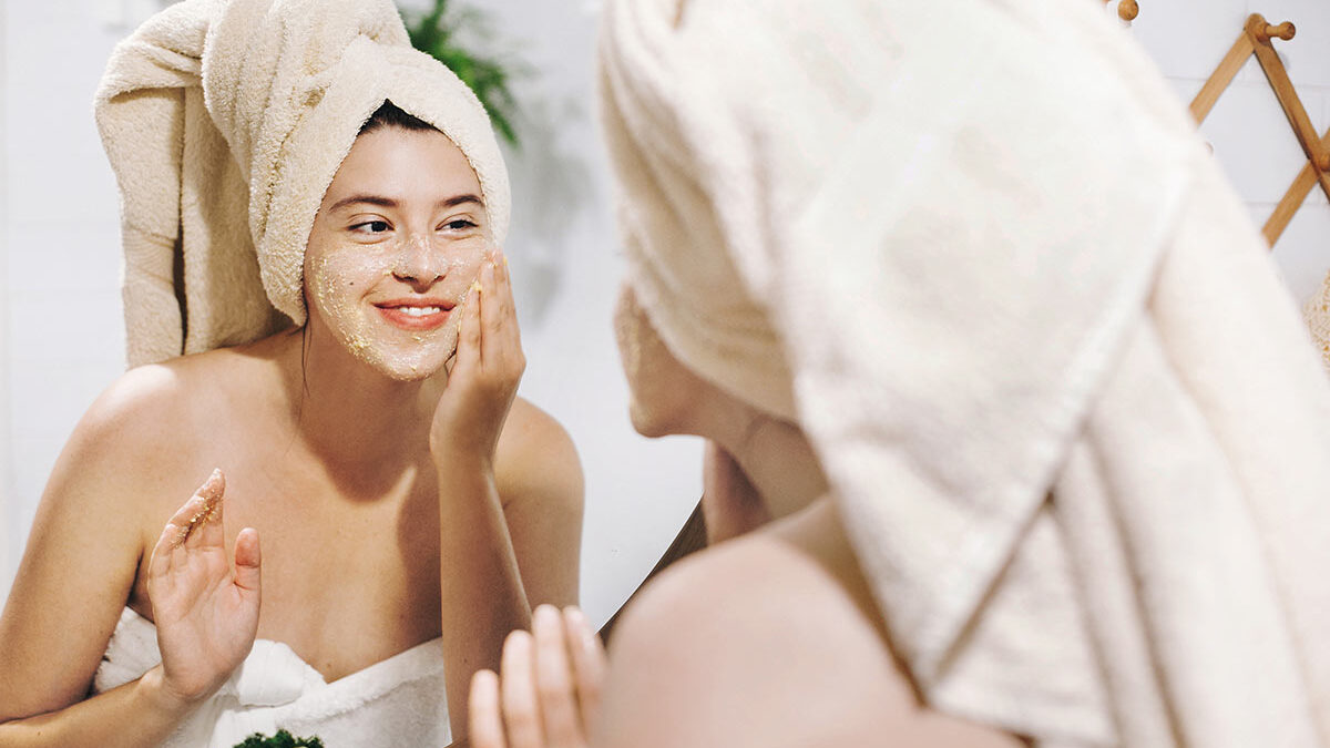 Mulher de toalha esfoliando a pele do rosto e se olhando no espelho